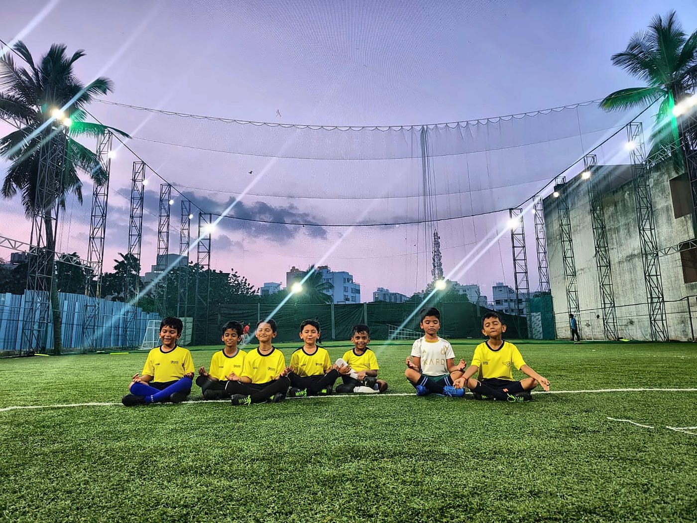 Young players sitting cross-legged on the turf at dusk under the floodlights